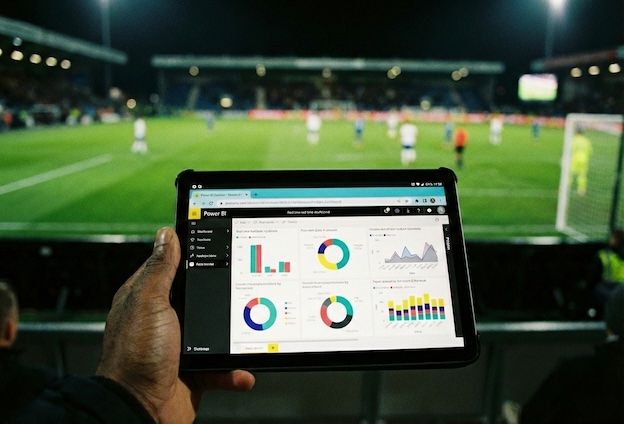 Em primeiro plano, uma mão segurando um Tablet rodando um Dashboard do Power BI (com gráficos coloridos), e ao fundo (desfocado), o campo de futebol iluminado à noite.